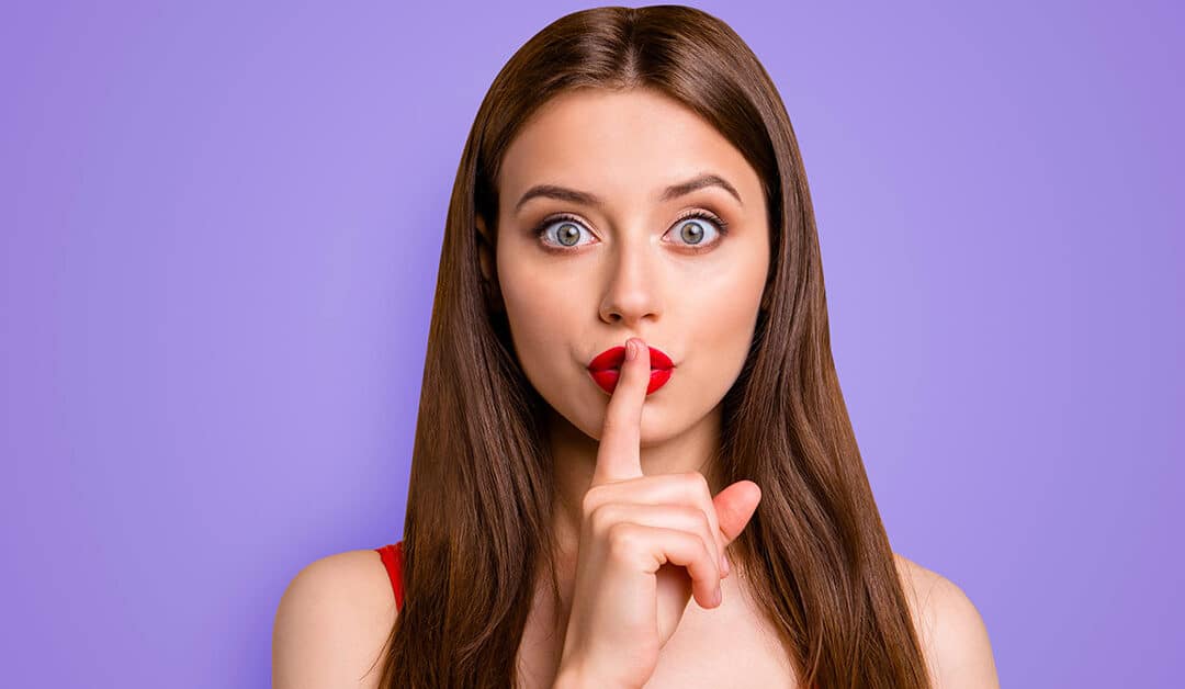 ein junge Frau, die sich den Zeigefinger vor den Mund hält um Verschwiegenheit zu demonstrieren