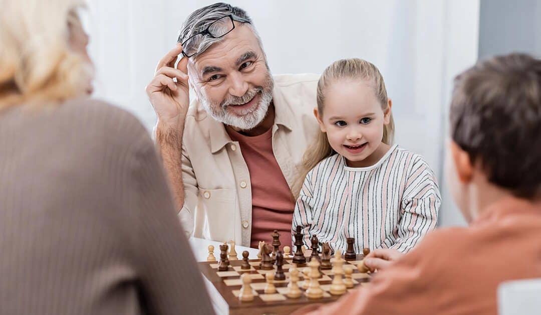 Ein Seniorenpärchen sitzt mit seinen Enkelkindern glücklich im Wohnzimmer und spielt Schach als Symbolbild für strategisch kluge Entscheidungen bei Investments für die Altersvorsorge | Wohnen im Alter